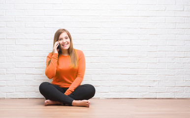 Young adult woman sitting on the floor over white brick wall calling using smartphone with a happy...