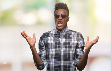 Naklejka premium Young african american man wearing sunglasses over isolated background crazy and mad shouting and yelling with aggressive expression and arms raised. Frustration concept.