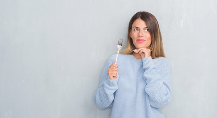 Young adult woman over grey grunge wall holding silver fork serious face thinking about question,...