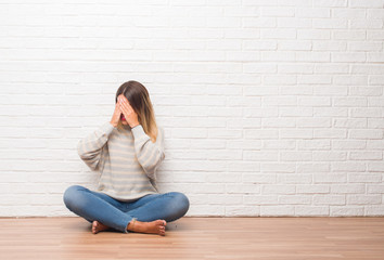 Fototapeta na wymiar Young adult woman sitting on the floor over white brick wall at home with sad expression covering face with hands while crying. Depression concept.