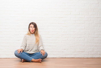 Young adult woman sitting on the floor over white brick wall at home making fish face with lips, crazy and comical gesture. Funny expression.