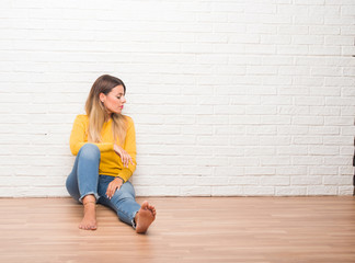 Young adult woman sitting on the floor over white brick wall looking to side, relax profile pose with natural face with confident smile.