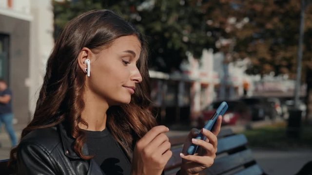 Portrait of a beautiful young modern brunette woman, in a leather jacket with airpods in her ear, listens to music. girl smiling, looking down and take call happy with phone.