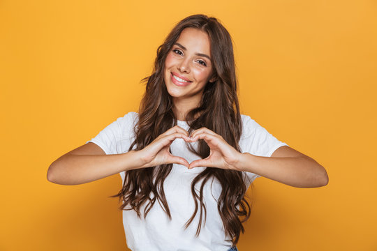 Image of charming woman 20s with long hair smiling and showing heart shape with fingers, isolated over yellow background
