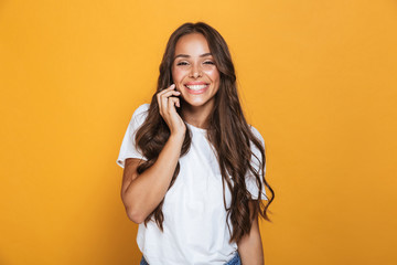 Portrait of charming woman 20s with long hair smiling and talking on mobile phone, isolated over yellow background