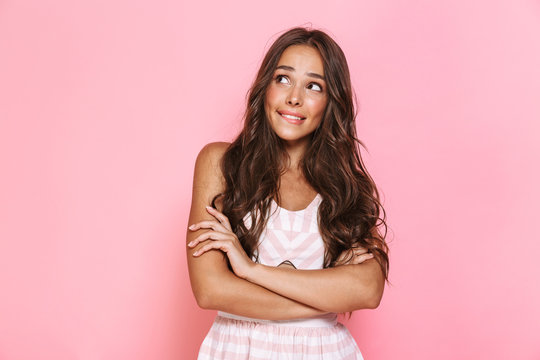 Image of charming woman 20s with long hair wearing dress smiling aside with arms crossed, isolated over pink background