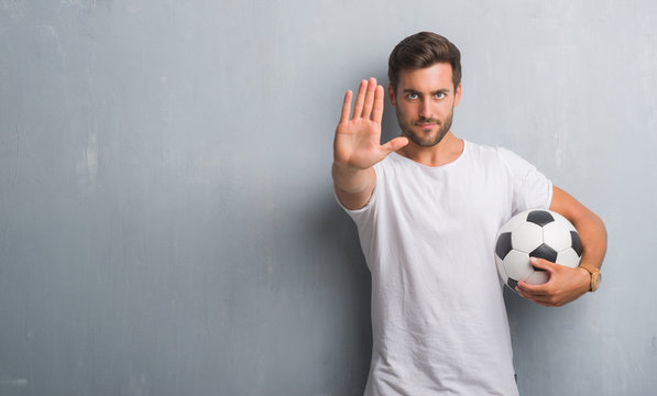 Handsome Young Man Over Grey Grunge Wall Holding Soccer Football Ball With Open Hand Doing Stop Sign With Serious And Confident Expression, Defense Gesture