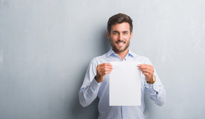 Handsome young man over grey grunge wall holding blank paper sheet contract with a happy face...