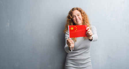 Young redhead woman over grey grunge wall holding flag of China with a happy face standing and smiling with a confident smile showing teeth