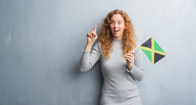 Young redhead woman over grey grunge wall holding flag of Jamaica surprised with an idea or question pointing finger with happy face, number one
