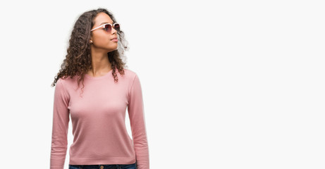 Beautiful young hispanic woman wearing sunglasses smiling looking side and staring away thinking.