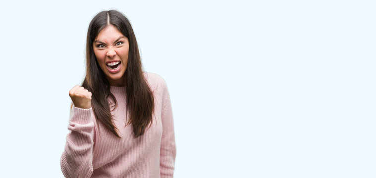 Young beautiful hispanic woman wearing a sweater angry and mad raising fist frustrated and furious while shouting with anger. Rage and aggressive concept.