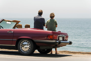 A middle aged couple with parked convertible car, looking at Pacific ocean near highway - obrazy, fototapety, plakaty