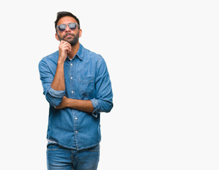 Fototapeta premium Adult hispanic man wearing sunglasses over isolated background with hand on chin thinking about question, pensive expression. Smiling with thoughtful face. Doubt concept.