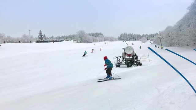 Child learns to ski and falls on skiing downhill on resort at winter day. Ski training on slope. Boy crash down on ski trail, people ride down from top. Winter sport with skier learning on piste