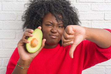 Young african american woman over white brick wall eating avocado with angry face, negative sign...