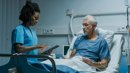 In the Hospital, Senior Patient Lying in the Bed Talking to a Nurse who is Holding Tablet Computer...