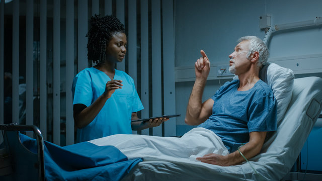 In the Hospital, Senior Patient Lying in the Bed Nurse Holding Tablet Computer Shows Augmented Reality Information, They Both Make Gestures and Talk. In the Technologically Advanced Hospital Ward.