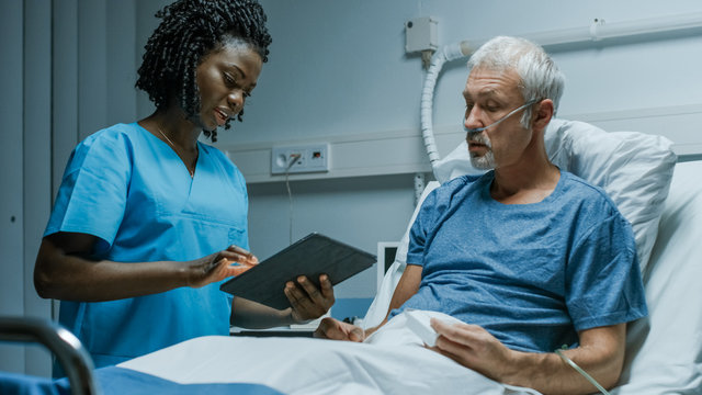 In the Hospital, Senior Patient Lying in the Bed Talking to a Nurse who is Holding Tablet Computer Showing Him Information. In the Technologically Advanced Hospital Ward.
