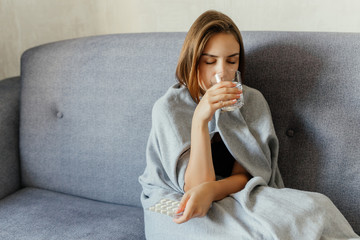 A woman wrapped in a grey blanket holding pills and drinking water, freezing, sitting on the sofa, at home.