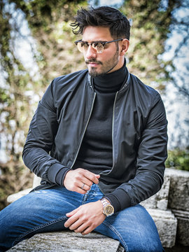 One handsome young man in urban setting in modern city, sitting, wearing black leather jacket and jeans