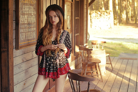 Girl in the Wild West, in Western house. Girl in hat with long cerly hair. Beautiful pretty girl in black hat. Incredible trip, travel. Cute woman on the terrace in red dress pouse.