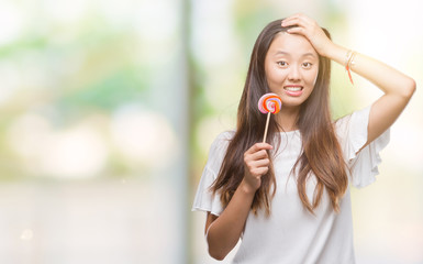 Young asian woman eating lollipop candy over isolated background stressed with hand on head,...