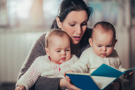 Charming mother showing images in a book to her cute twin babies at home
