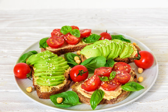 wholegrain bread avocado and tomatoes toasts with hummus, sesame and basil in a plate over white wooden table