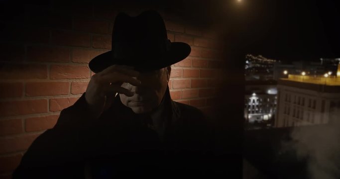 A classic film noir 1940s style scene with a suspicious looking character wearing a fedora lurking in the shadows of a dimly lit rooftop.