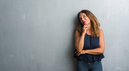 Naklejka premium Middle age hispanic woman standing over grey grunge wall with hand on chin thinking about question, pensive expression. Smiling with thoughtful face. Doubt concept.