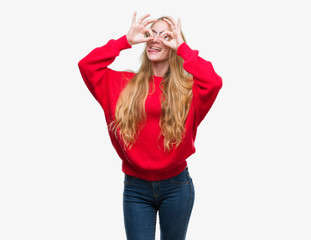 Obraz na płótnie Canvas Blonde teenager woman wearing red sweater doing ok gesture like binoculars sticking tongue out, eyes looking through fingers. Crazy expression.