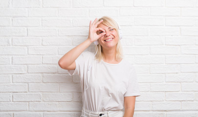 Adult caucasian woman over white brick wall wearing headphones doing ok gesture with hand smiling, eye looking through fingers with happy face.