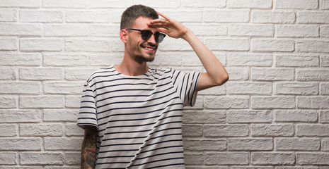 Young adult man wearing sunglasses standing over white brick wall very happy and smiling looking...