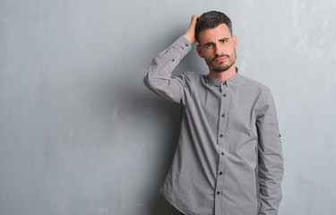 Young adult man standing over grey grunge wall confuse and wonder about question. Uncertain with doubt, thinking with hand on head. Pensive concept.