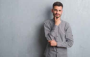 Young adult man standing over grey grunge wall looking confident at the camera with smile with crossed arms and hand raised on chin. Thinking positive.