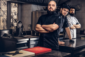 Team of professional bearded cooks dressed in uniforms standing in kitchen.