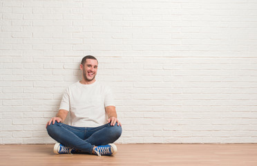 Obraz na płótnie Canvas Young caucasian man sitting on the floor over white brick wall sticking tongue out happy with funny expression. Emotion concept.