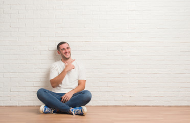 Young caucasian man sitting on the floor over white brick wall cheerful with a smile of face pointing with hand and finger up to the side with happy and natural expression on face
