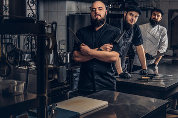 Team of professional bearded cooks dressed in uniforms posing with knives in kitchen.
