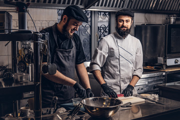 Two smiling bearded cooks dressed in uniforms preparing sushi in the kitchen.