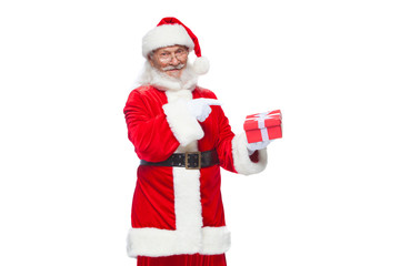 Obraz premium Christmas. Smiling Santa Claus in white gloves is holding a gift red box with a bow. Pointing at the gift. Isolated on white background.