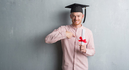 Young redhead man over grey grunge wall wearing graduate cap holding degree with surprise face pointing finger to himself