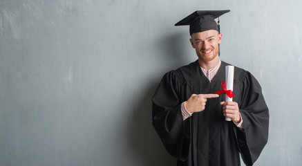 Young redhead man over grey grunge wall wearing graduate uniform holding degree very happy pointing...