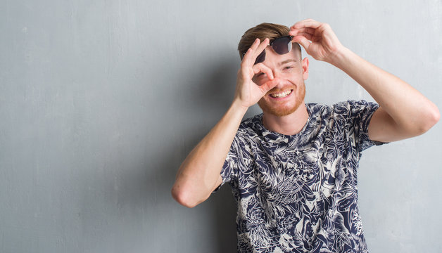 Young redhead man over grey grunge wall wearing sunglasses with happy face smiling doing ok sign with hand on eye looking through fingers