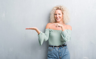 Young blonde woman with curly hair over grunge grey background amazed and smiling to the camera while presenting with hand and pointing with finger.