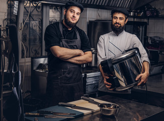Two brutal cooks dressed in uniforms and hats standing with big multicooker in the kitchen.