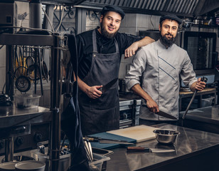 Two bearded cooks dressed in uniforms and hats posing in kitchen, looking at a camera.