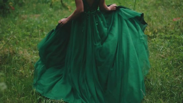 Red-haired girl in a luxurious green dress with sparkling beads. The princess runs through a forest glade, a long train waving in the wind. Shooting from the back without a face. Slow motion.
