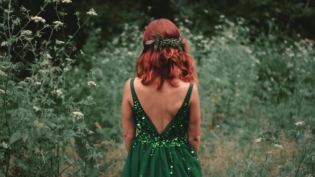 Red-haired girl a forest nymph in a luxurious, long, green dress, which is embroidered with sparkling beads. The princess slowly leaves the dark forest thicket. The image of a halloween party